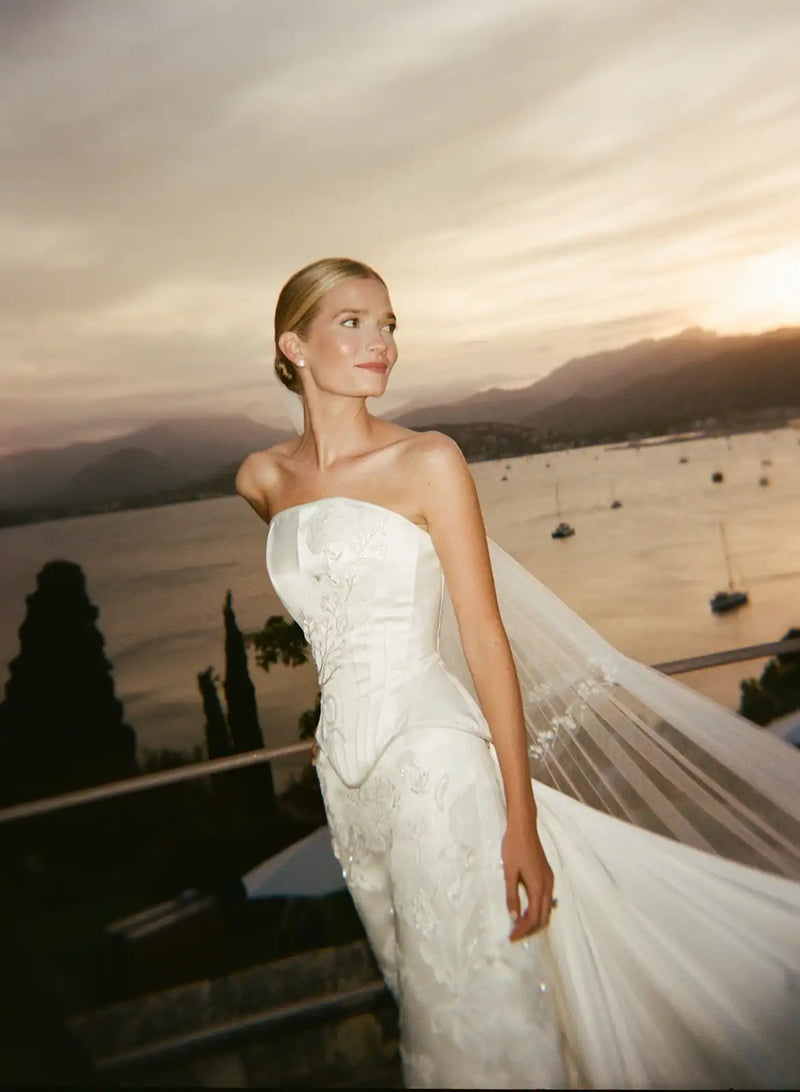 Woman in a white wedding dress standing by a scenic waterfront at sunset.