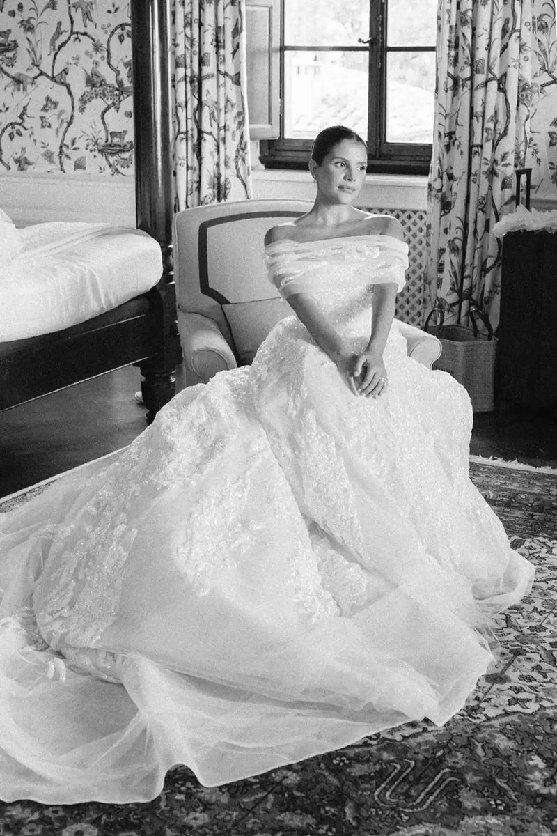 Woman in a white wedding dress sitting in a room with patterned curtains and furniture.