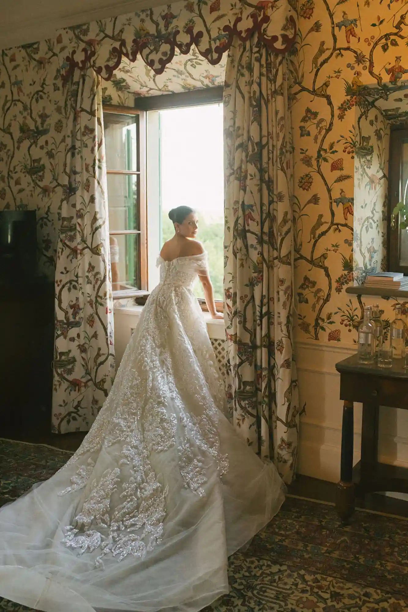Woman in a white lace wedding dress standing in a room with floral wallpaper.