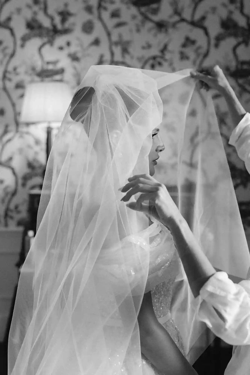 Black and white photo of a woman wearing a wedding veil with a decorative wall in the background.