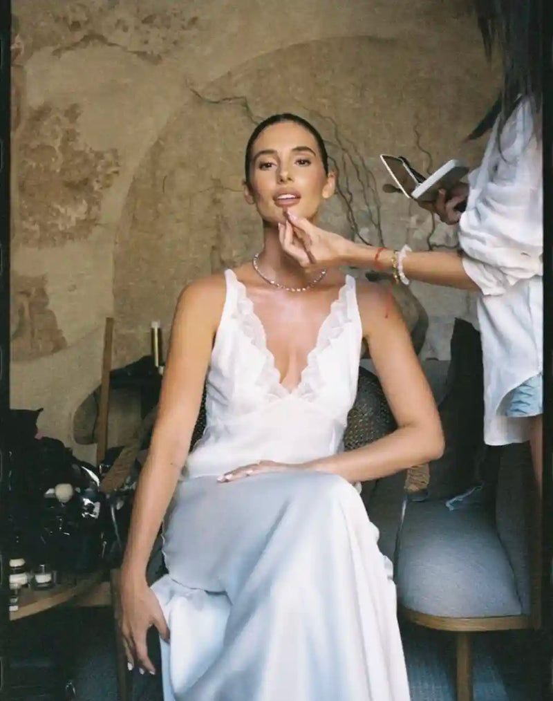 Woman in a white dress being made up by a makeup artist in a rustic setting.