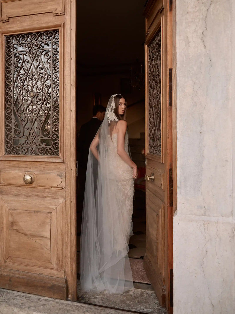Woman in a wedding dress and veil exiting a wooden door with intricate designs.