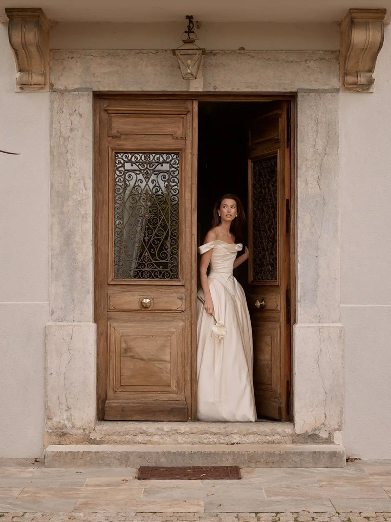 Woman in a white off-shoulder dress standing in an open wooden door of a stone building.