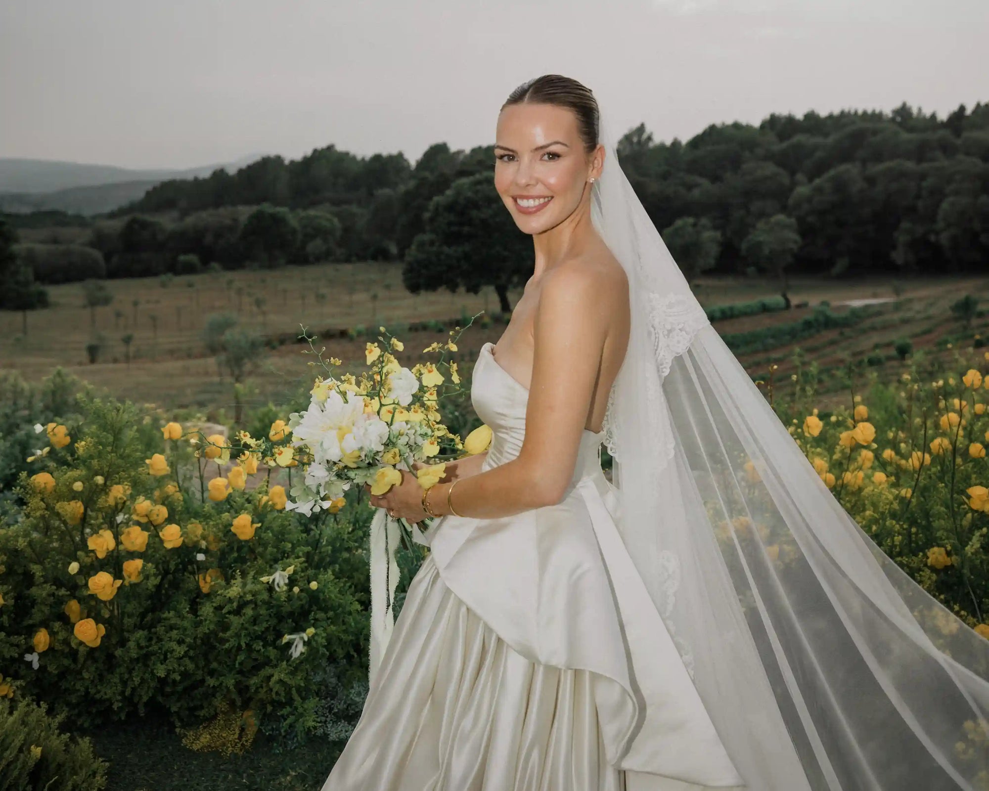 Bride holding a bouquet of flowers in a scenic outdoor setting with trees and fields.