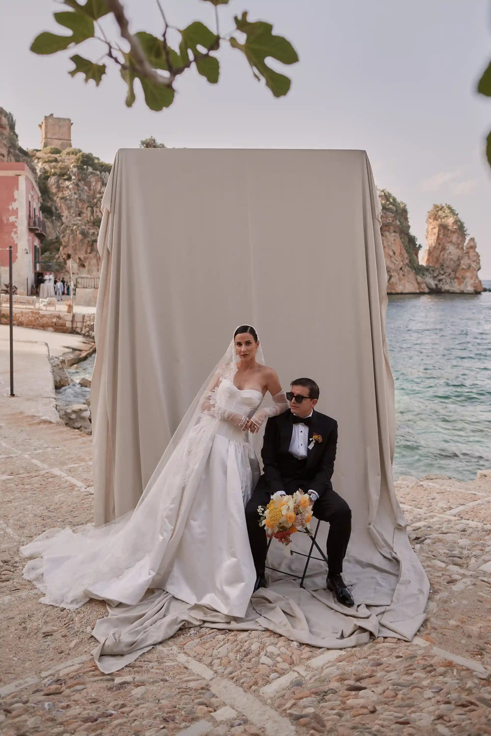 Wedding couple posing under a draped canopy with a scenic coastal background.