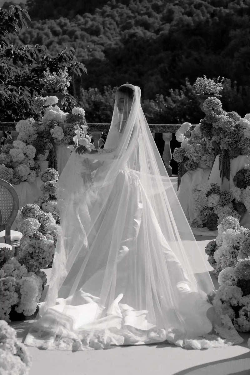 Bride in a white wedding dress with a long veil standing in front of floral decorations.