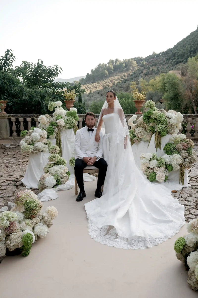Wedding couple sitting on a bench with floral arrangements in an outdoor setting.